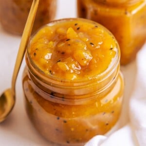 A jar full to the brim of mango chutney with a brass coloured spoon leaning up against the jar.