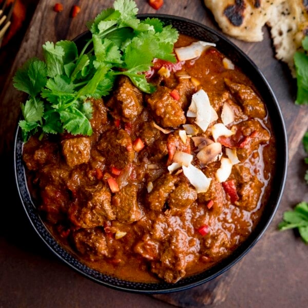 Square image of beef curry, topped with coriander, chillies and toasted coconut on a black bowl. The bowl is on a dark brown surface and there are some ingredients scattered around the bowl, just in shot.