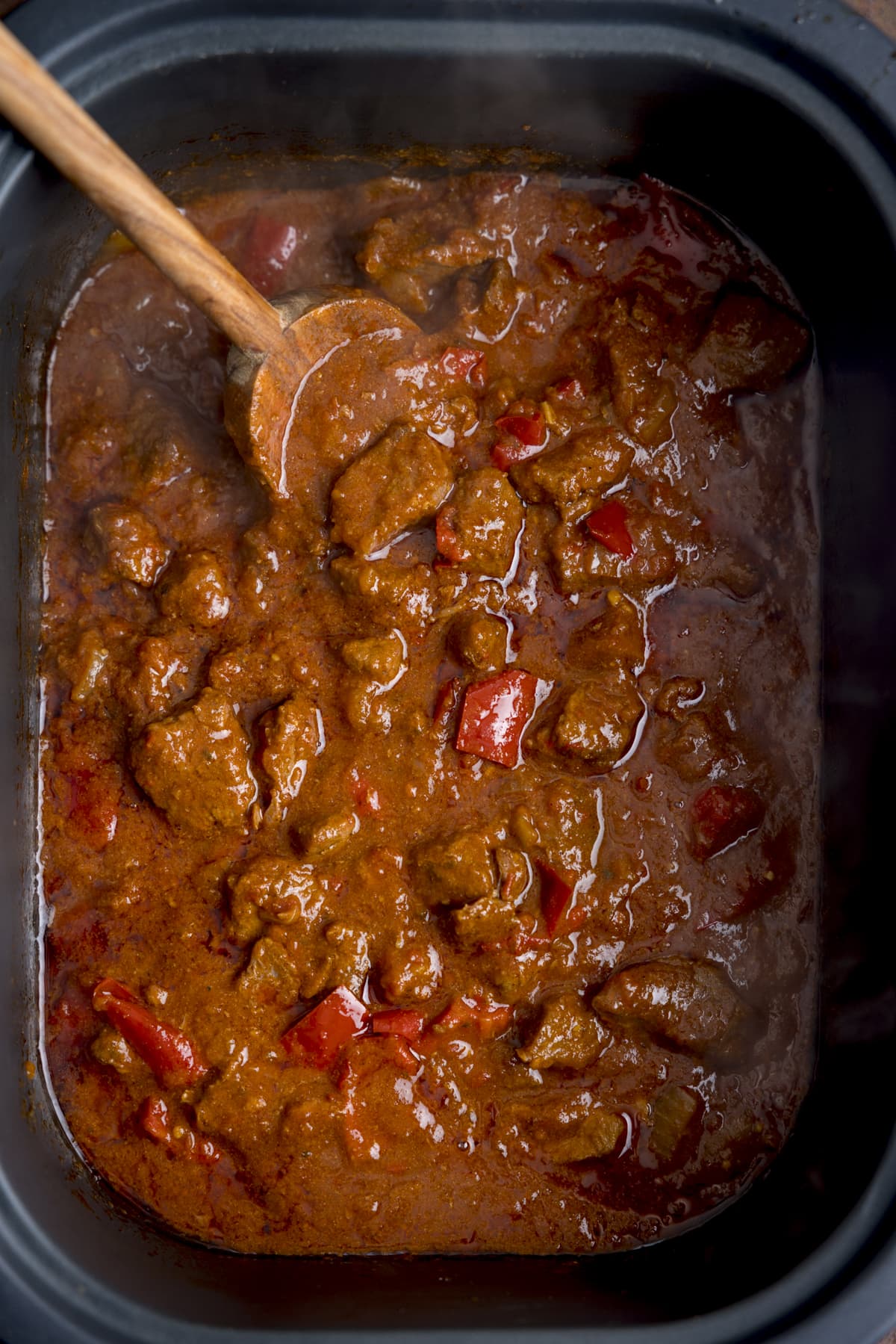 Overhead image of beef curry with red peppers in a slow cooker. There is a wooden spoon in the curry.