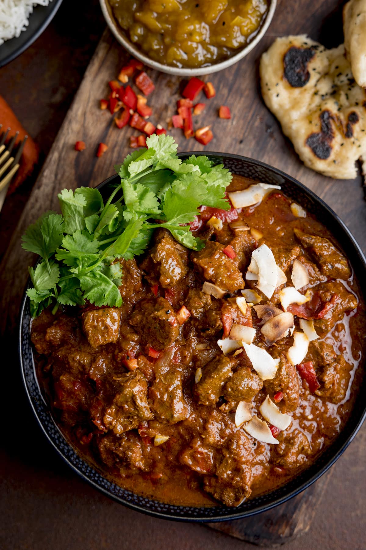 Overhead image of slow cooker beef curry in a black bowl, topped with coriander and toasted coconut. The bowl is on a wooden board and there is naan bread, chopped chillies and mango chutney around the dish