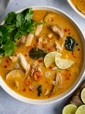 Tom Kha Gai - Thai chicken soup with coconut milk and galangal - in a white bowl on a light background. The soup is garnished with lime slices and coriander.