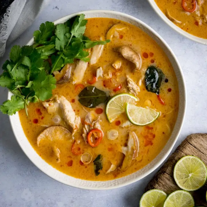 Tom Kha Gai - Thai chicken soup with coconut milk and galangal - in a white bowl on a light background. The soup is garnished with lime slices and coriander.