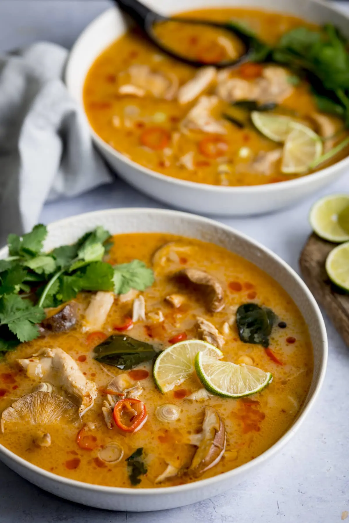 Tom Kha Gai - Thai chicken soup with coconut milk and galangal - in a white bowl on a light background. The soup is garnished with lime slices and coriander. There is a second bowl of soup in the background with a black spoon in it.