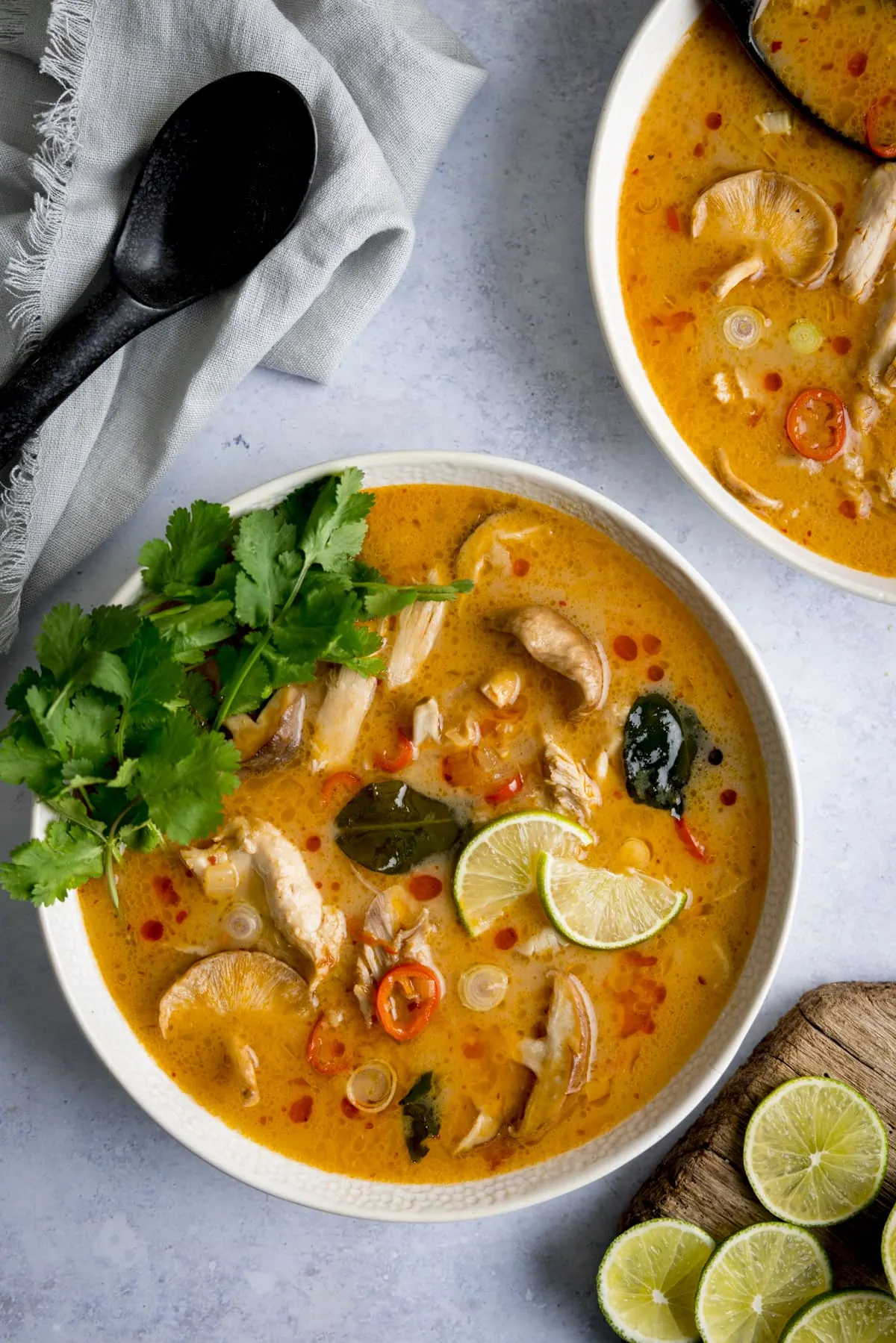 Tom Kha Gai - Thai chicken soup with coconut milk and galangal - in a white bowl on a light background. The soup is garnished with lime slices and coriander. There is a spoon, napkin and further slices of lime around the bowl. A second bowl of soup is just in shot.