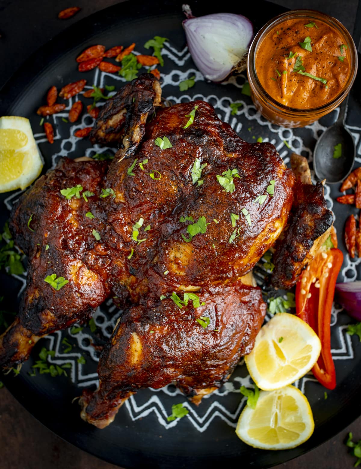 Spatch-cocked peri peri roast chicken on a black and white tray, surrounded by garnishes and a jar of homemade peri peri sauce.