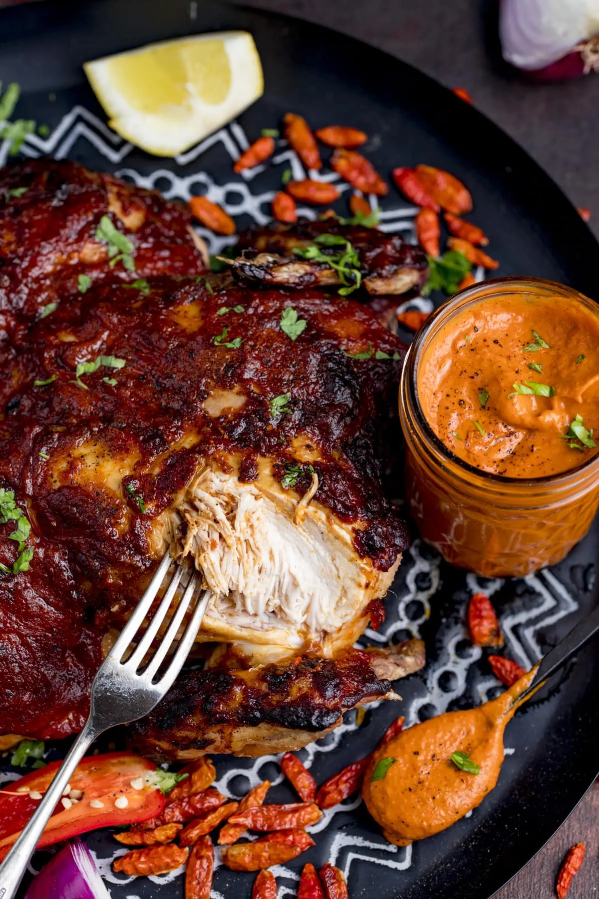 Peri peri roasted chicken on a black and white tray, next to a glass jar of homemade peri peri sauce. Part of the chicken is being shredded with a fork.