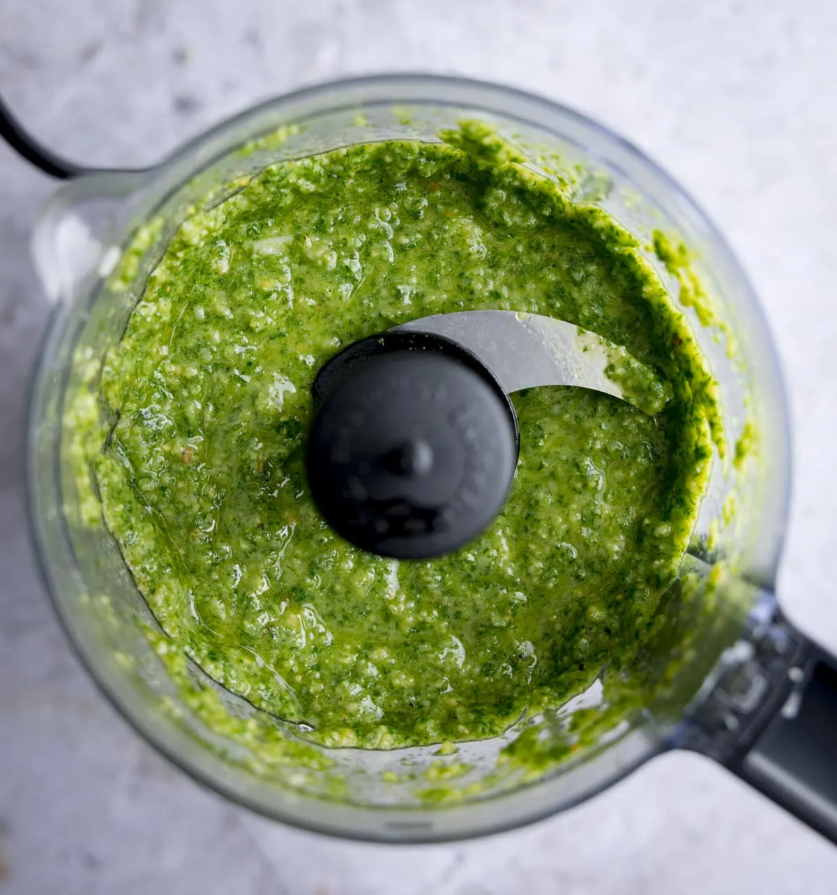 Overhead shot of homemade pesto in a small food processor, on a light background.