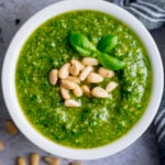 Overhead image of homemade pesto in a small white bowl, topped with pine nuts and basil leaves.