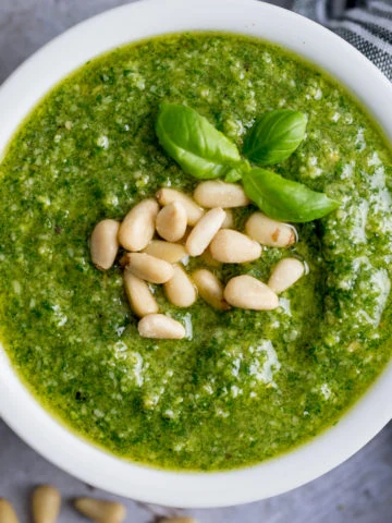 Overhead image of homemade pesto in a small white bowl, topped with pine nuts and basil leaves.
