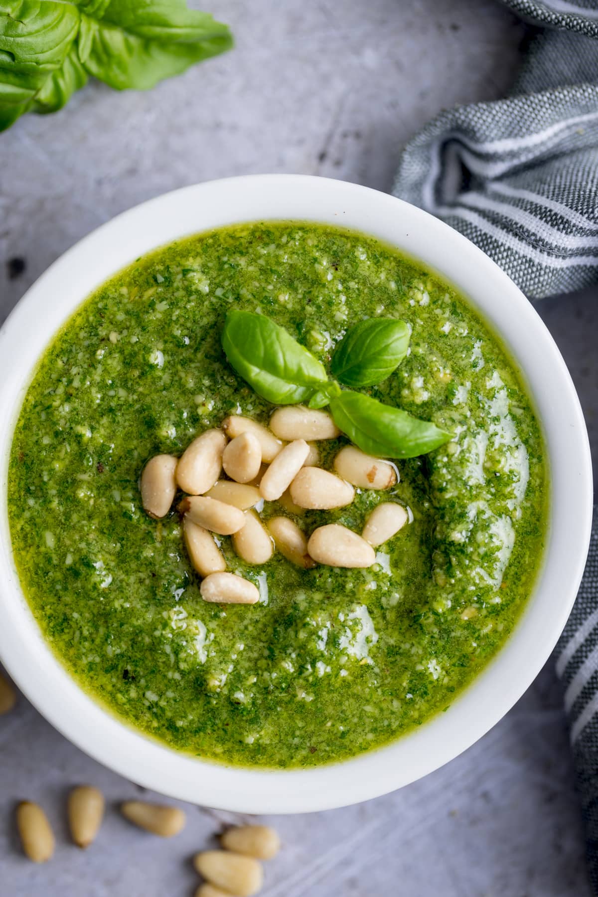 Pesto topped with pine nuts and basil in a small white bowl, surrounded by ingredients.