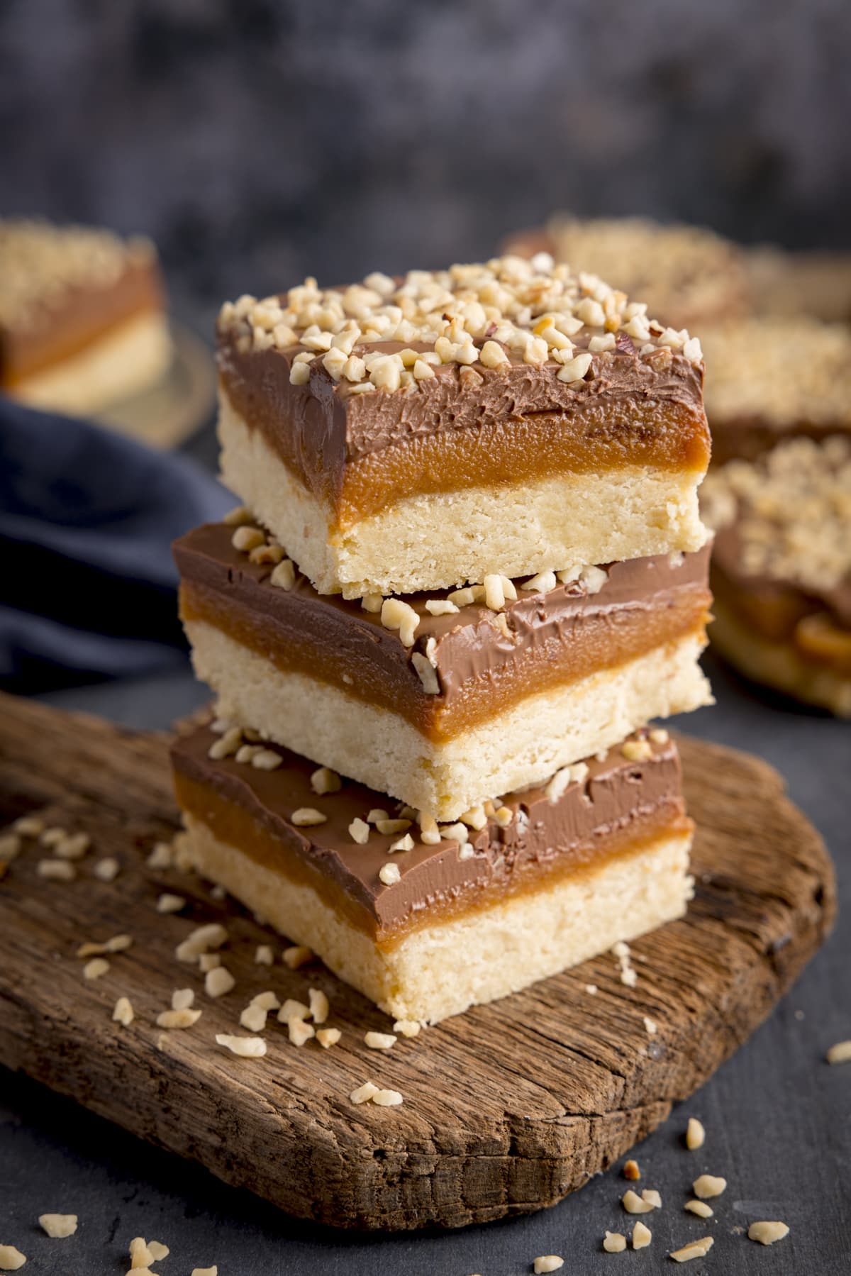 Three pieces of millionaire's shortbread, topped with chopped hazelnuts, piled up on a small rustic wooden board. The board is on a dark background, and there are further millionaire's shortbreads in the background.