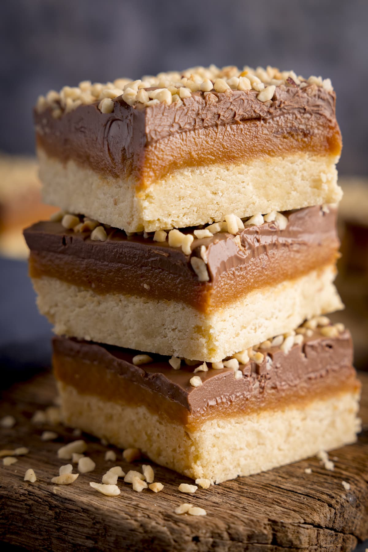 Extreme close-up of three pieces of millionaire's shortbread, topped with chopped hazelnuts, piled up on a small rustic wooden board. 