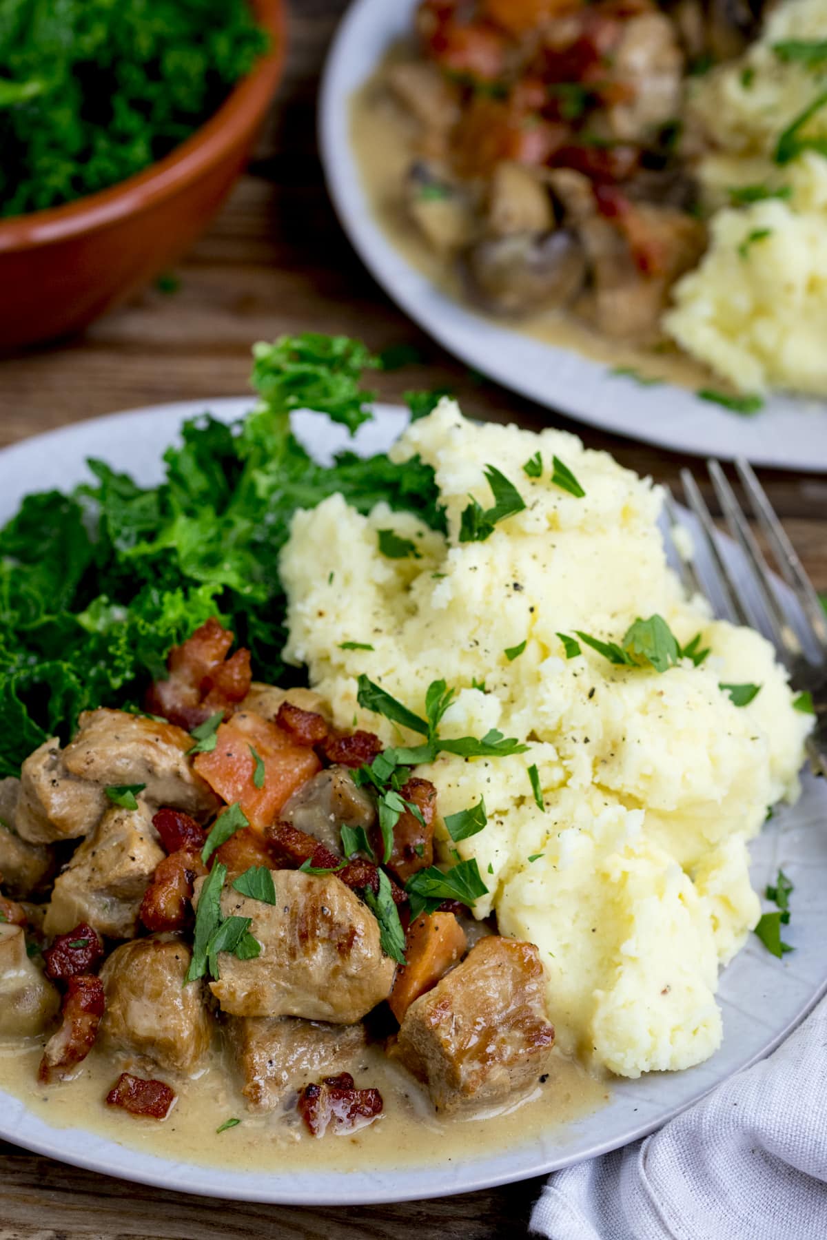 Creamy pork casserole, topped with bacon on a plate with mashed potato and kale. There is a further plate and a bowl of kale in the background.