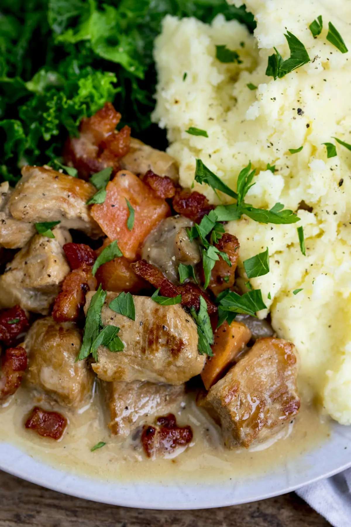 Close-up overhead image of cream pork casserole with bacon, carrot and mushroom on a light plate with mashed potato and kale. Parsley has been sprinkled on top.