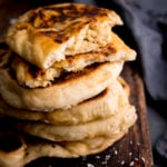 Close up of a stack of peshwari naan breads on a dark wooden board against a blue background. There is a blue napkin in shot. The top naan has been broken into two to show the filling.