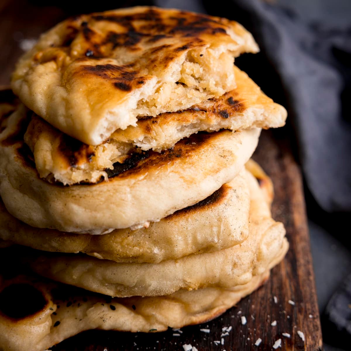 Close up of a stack of peshwari naan breads on a dark wooden board against a blue background. There is a blue napkin in shot. The top naan has been broken into two to show the filling.