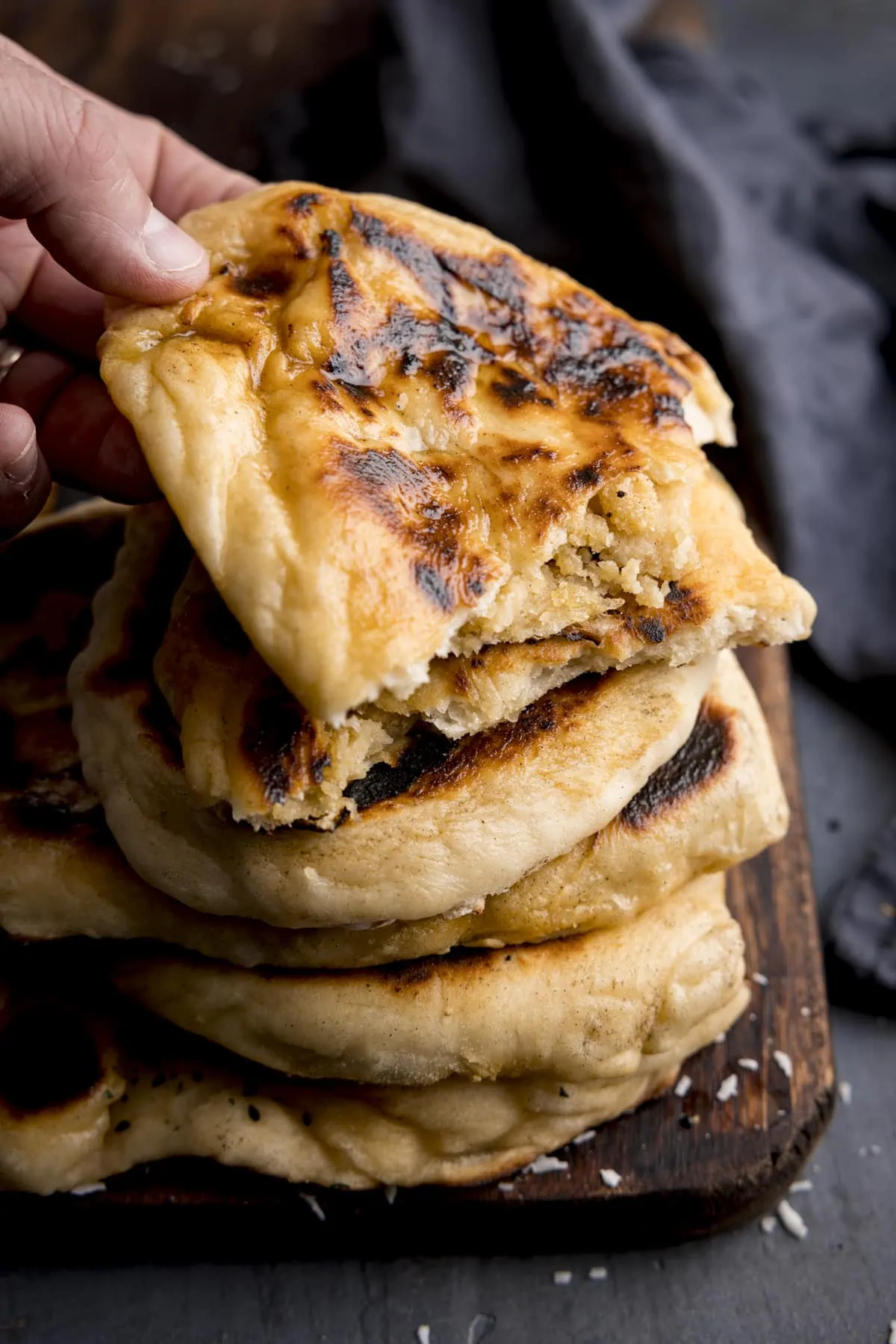 Tall image of a stack of peshwari naan breads on a wooden board against a blue background. There is a blue napkin in shot and a hand is taking the top naan.