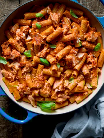 Square image of rigatoni and salmon in creamy tomato sauce in a blue cast iron pan on a wooden table.