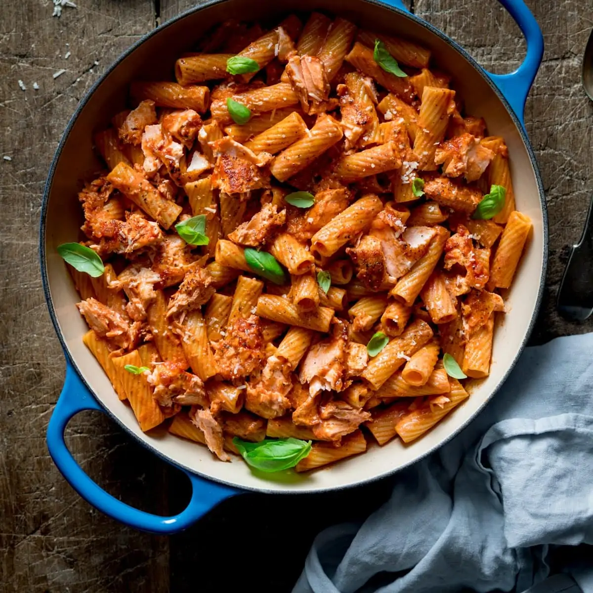 Square image of rigatoni and salmon in creamy tomato sauce in a blue cast iron pan on a wooden table.