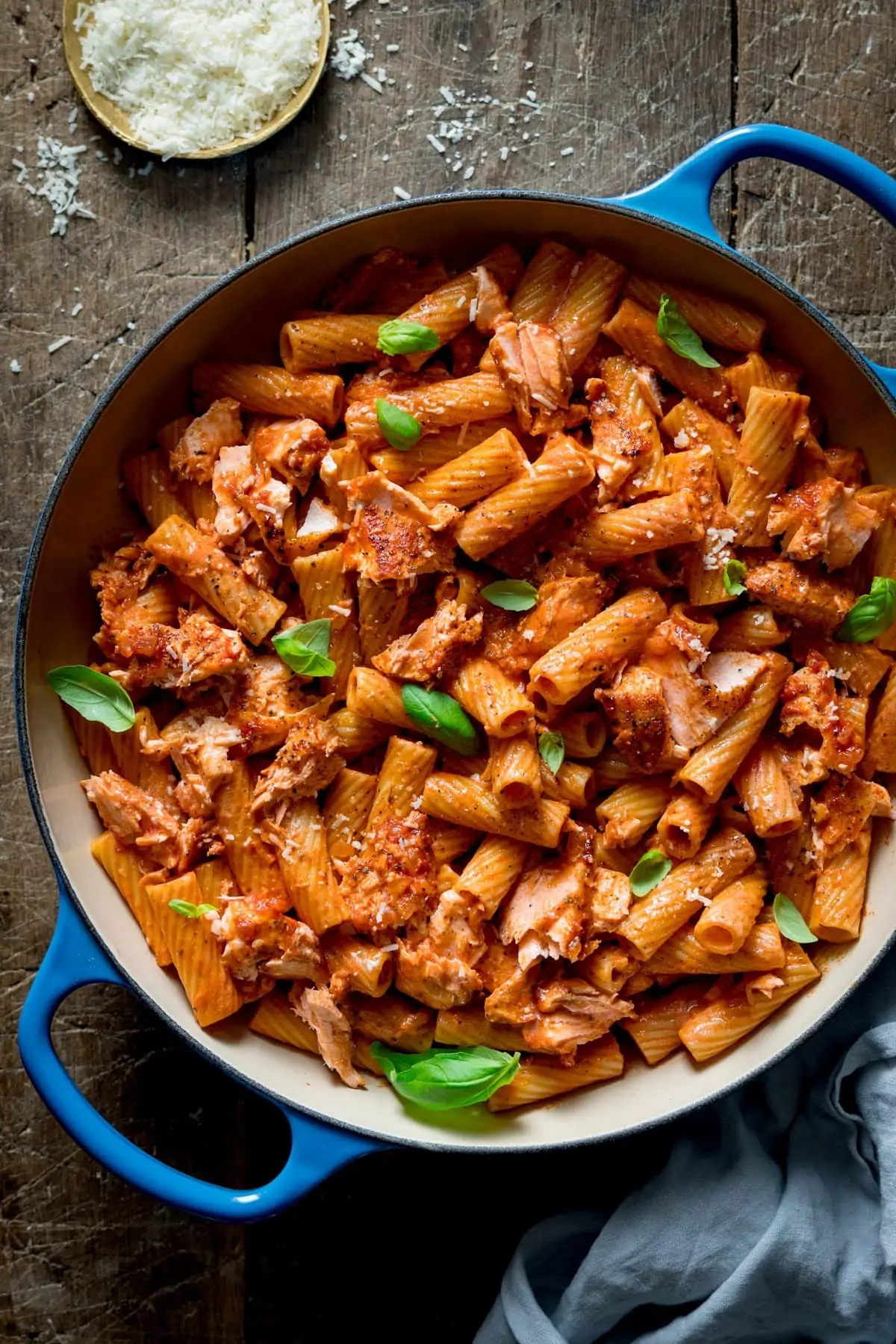 Overhead image of creamy tomato salmon and rigatoni in a blue cast iron pan on a wooden table. There is a dish of grated parmesan at the top of the image.