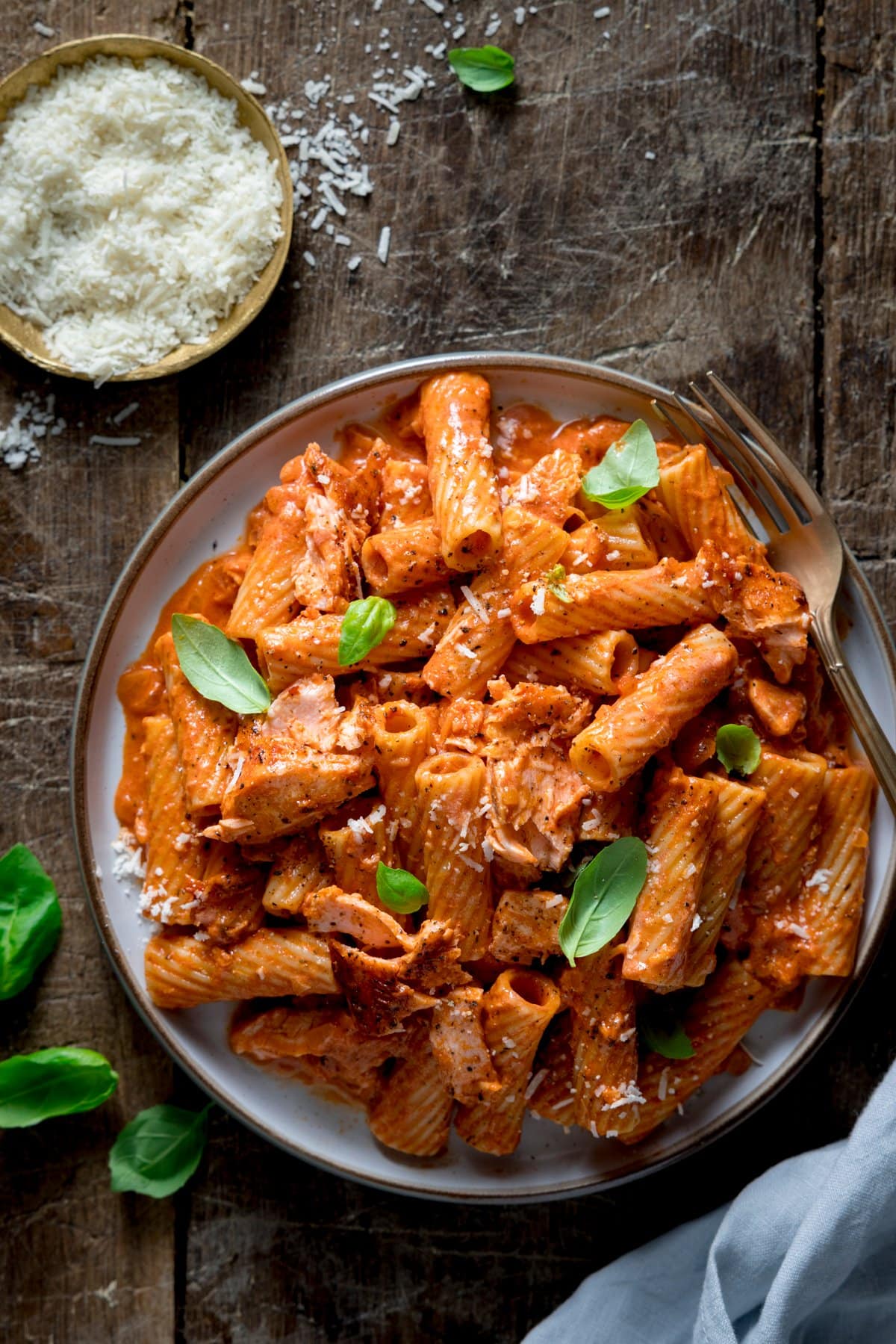 Overhead image of a plate of creamy tomato salmon pasta on a wooden table. There are basil leaves and a dish of grated parmesan around the plate.