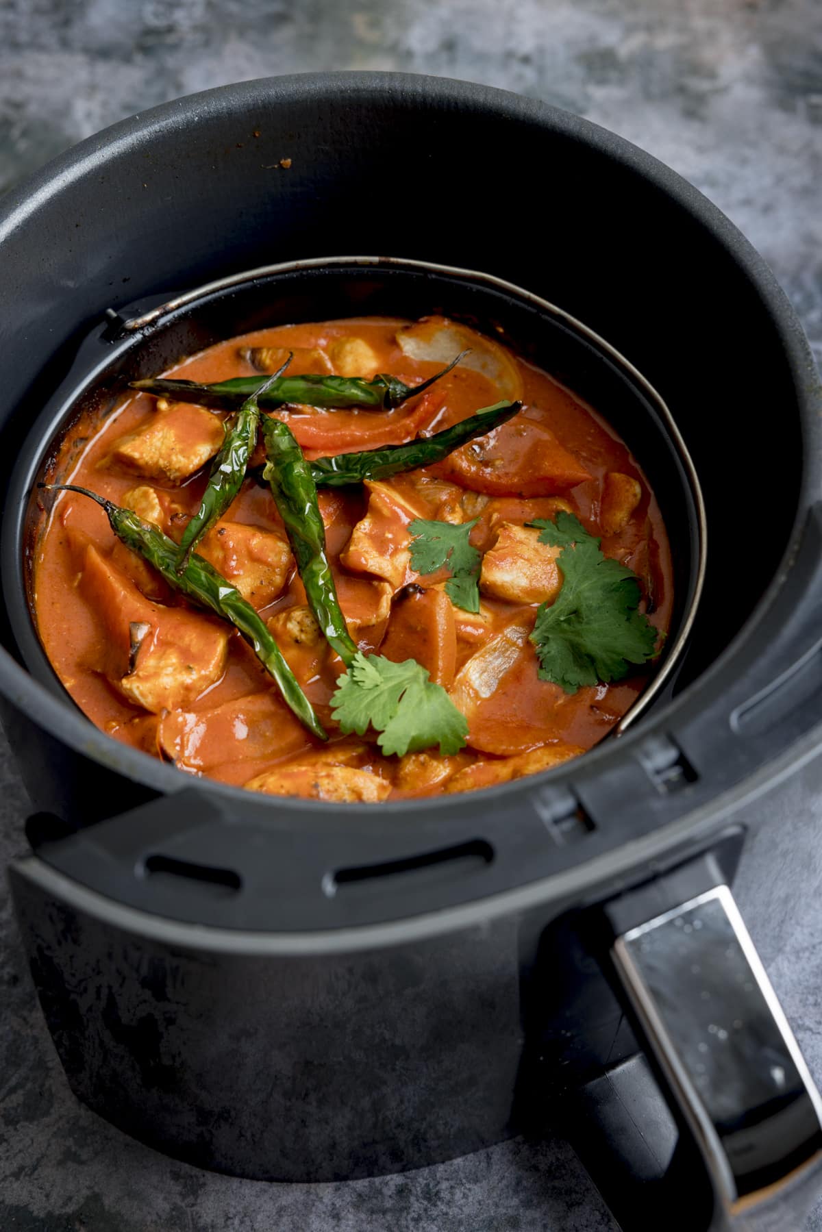 Tall image of Air Fryer Chicken Curry sprinkled with coriander and whole fried green chillies, inside a black air fryer with a stainless steel handle on a mottle grey work surface.