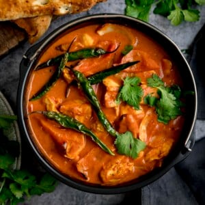 A square overhead shot of Air Fryer Chicken Curry sprinkled with coriander and whole fried green chillies, with bunches of coriander scattered around, there is a pile of toasted naan breads on a wooden board, some dark fabric napkins are to the side, against a mottle grey work surface.