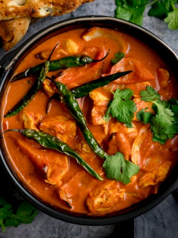 A square overhead shot of Air Fryer Chicken Curry sprinkled with coriander and whole fried green chillies, with bunches of coriander scattered around, there is a pile of toasted naan breads on a wooden board, some dark fabric napkins are to the side, against a mottle grey work surface.