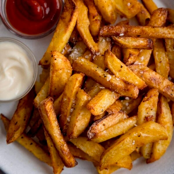 A square overhead shot of Air Fryer Chips sprinkled with salt and black pepper, on a white plate with a small bowl of ketchup on the side and a small bowl of mayonnaise.