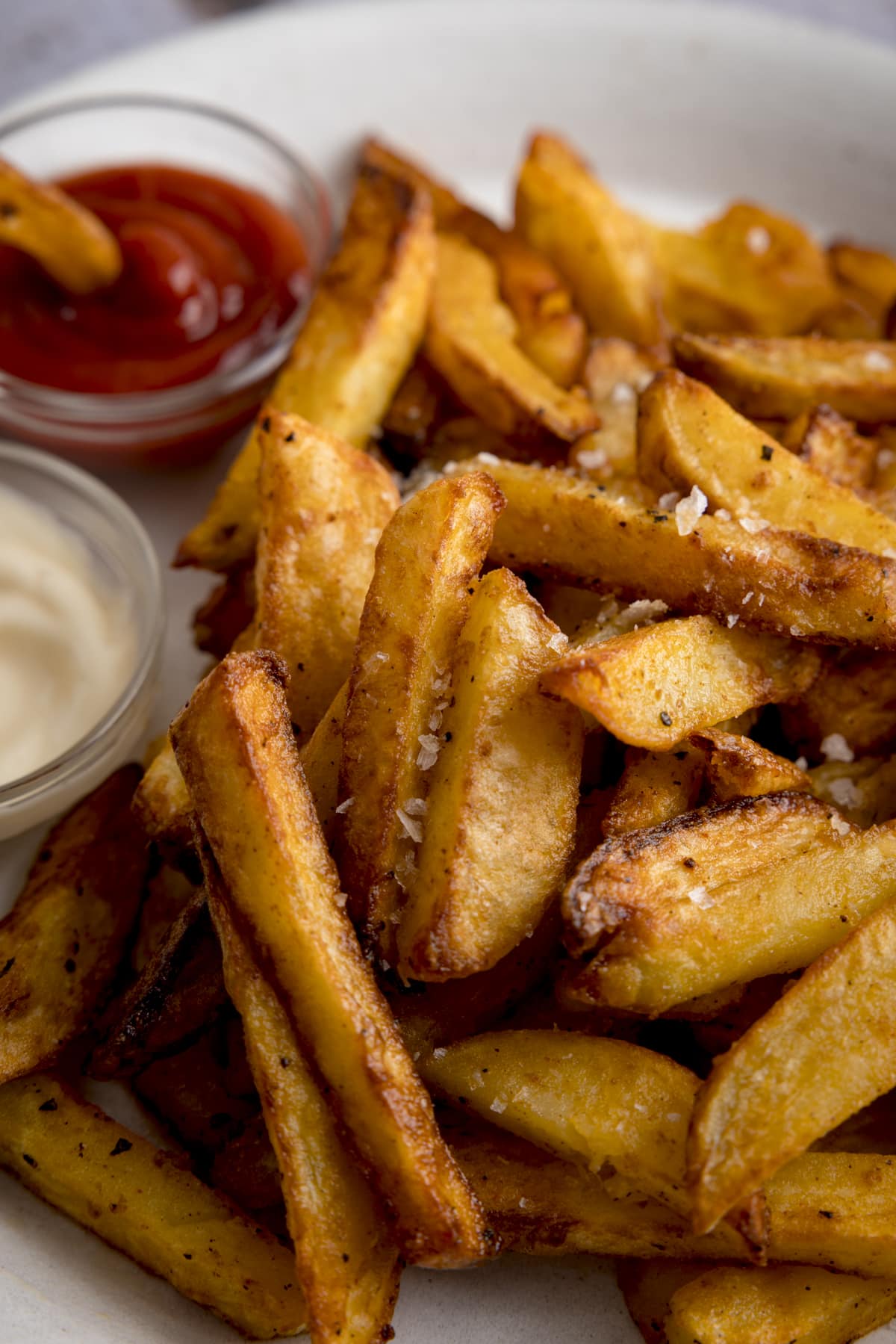 Close up of a mound of Air Fryer Chips sprinkled with salt and black pepper, on a white plate with a small bowl of ketchup with a chip dunked in on the side and a small bowl of mayonnaise.