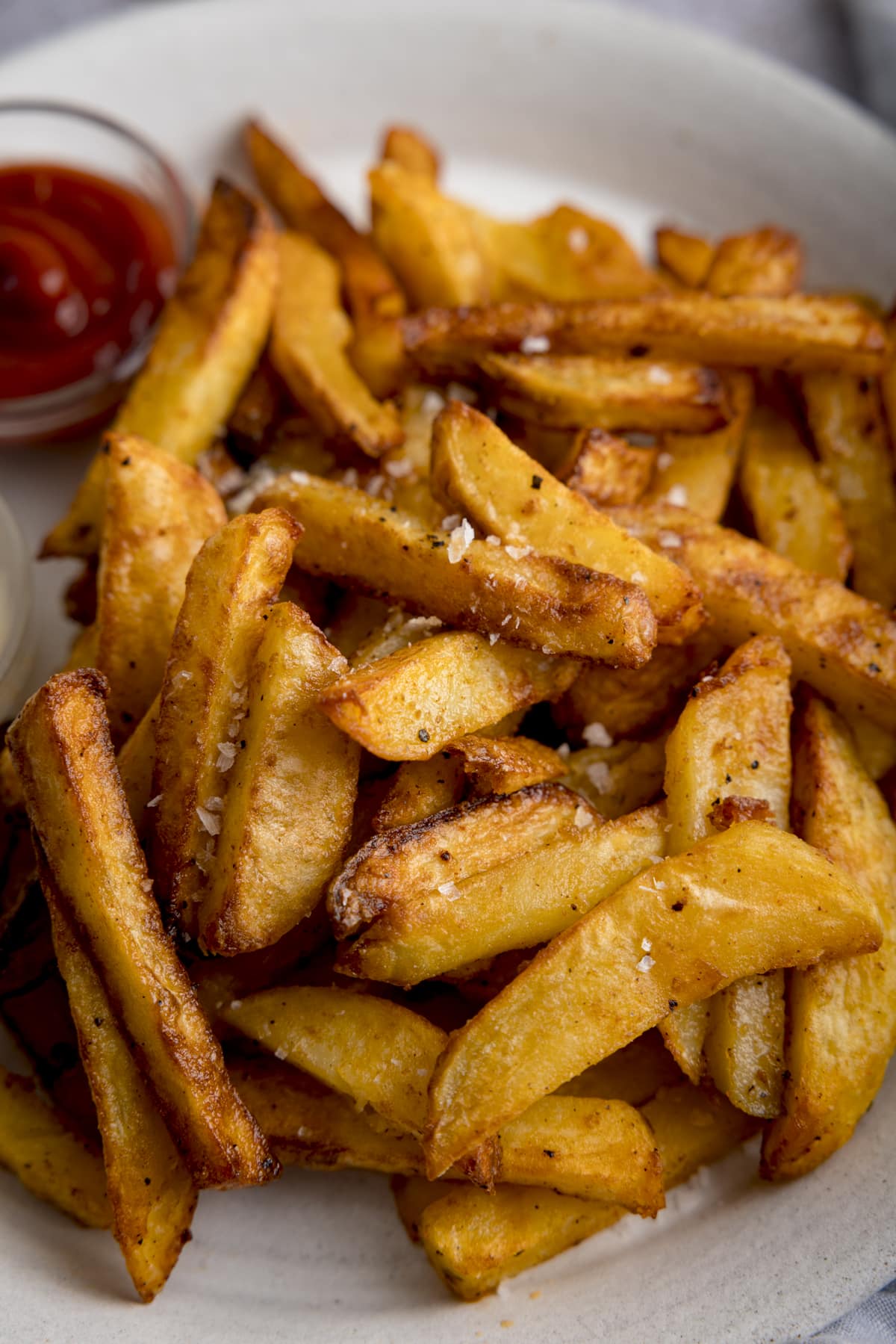 A tall close up shot of a mound of Air Fryer Chips sprinkled with salt and black pepper, on a white plate with a small bowl of ketchup on the side and a small bowl of mayonnaise.