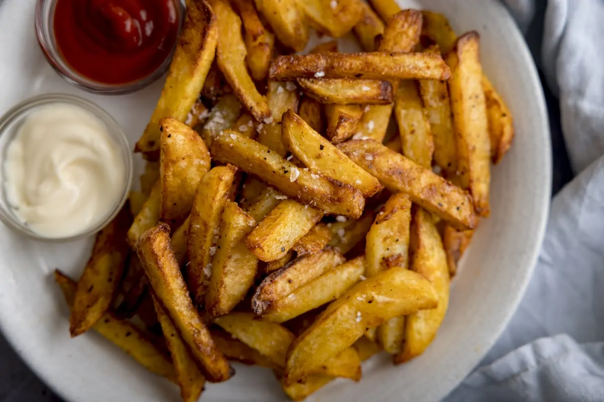 A wide close up shot of Air Fryer Chips sprinkled with salt and black pepper, on a white plate with a small bowl of ketchup on the side and a small bowl of mayonnaise.