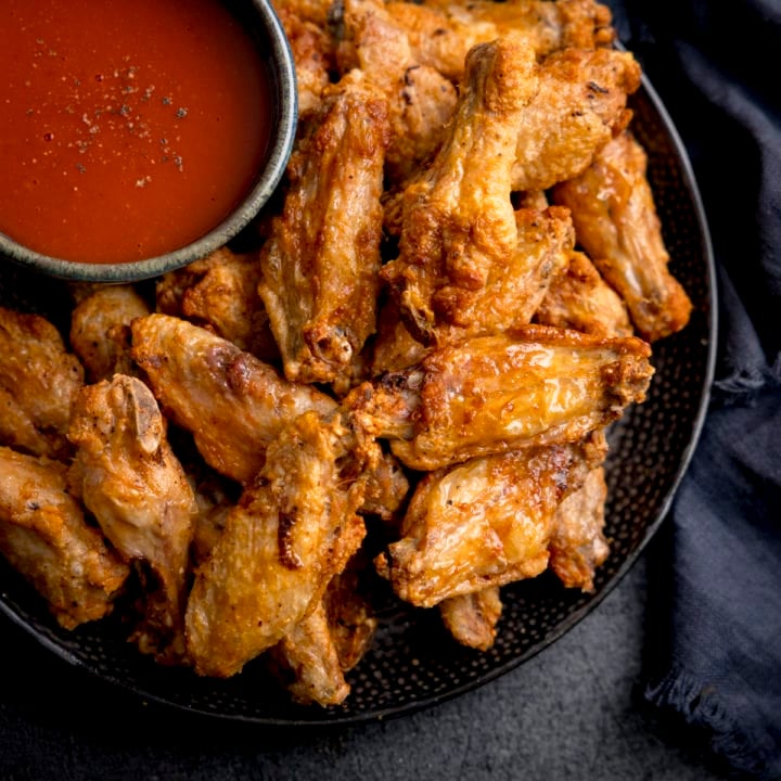 A square overhead shot of Air Fryer Crispy Chicken Wings piled up on a black speckled plate, their is also a large black bowl filled with buffalo sauce, sprinkled with seasoning on the plate, the plate is on a mottled dark grey background and there is some dark blue napkins to the side of the plate.