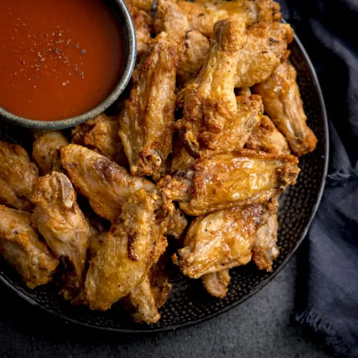 A square overhead shot of Air Fryer Crispy Chicken Wings piled up on a black speckled plate, their is also a large black bowl filled with buffalo sauce, sprinkled with seasoning on the plate, the plate is on a mottled dark grey background and there is some dark blue napkins to the side of the plate.
