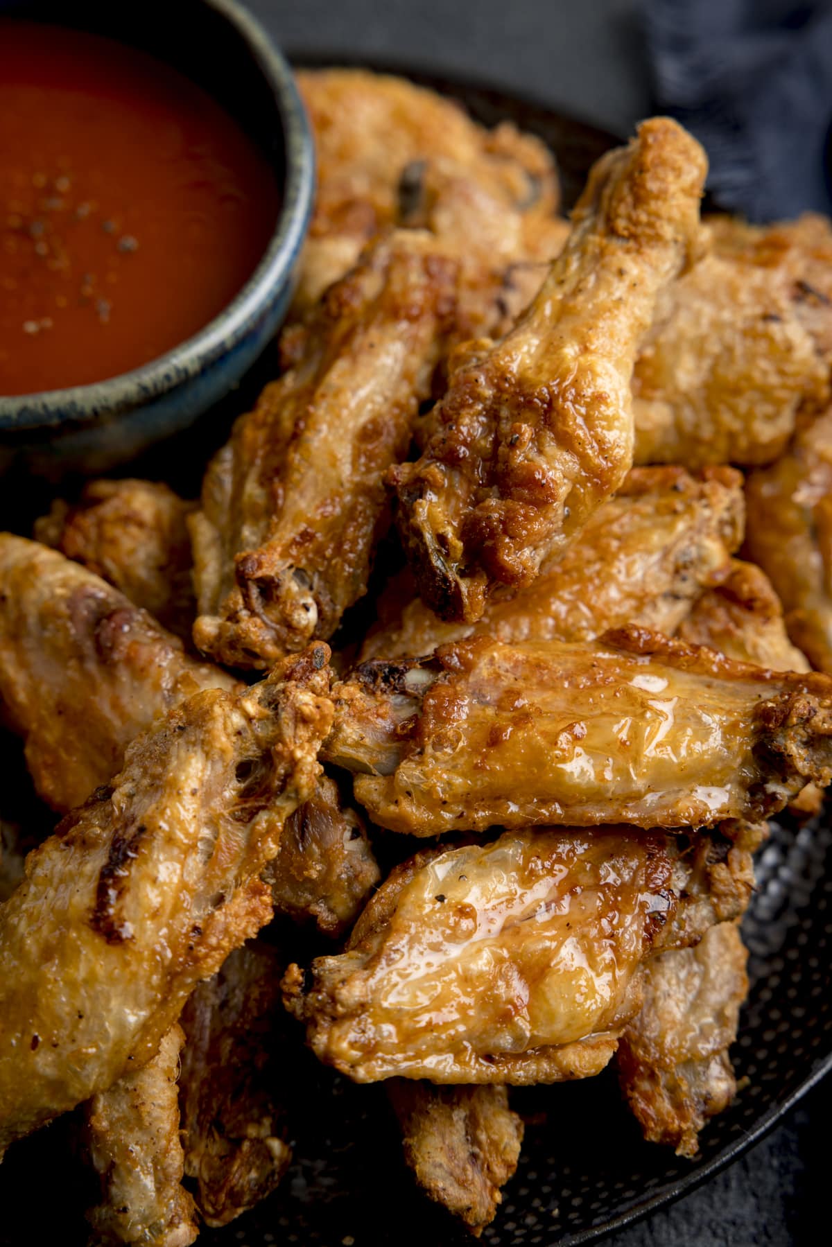 A closeup of Air Fryer Crispy Chicken Wings stacked on a black mottled plate, there is also a large black bowl of buffalo sauce sprinkled with spices on the plate, the plate is on a mottled dark gray background and there is a large one dark blue napkin in the background.