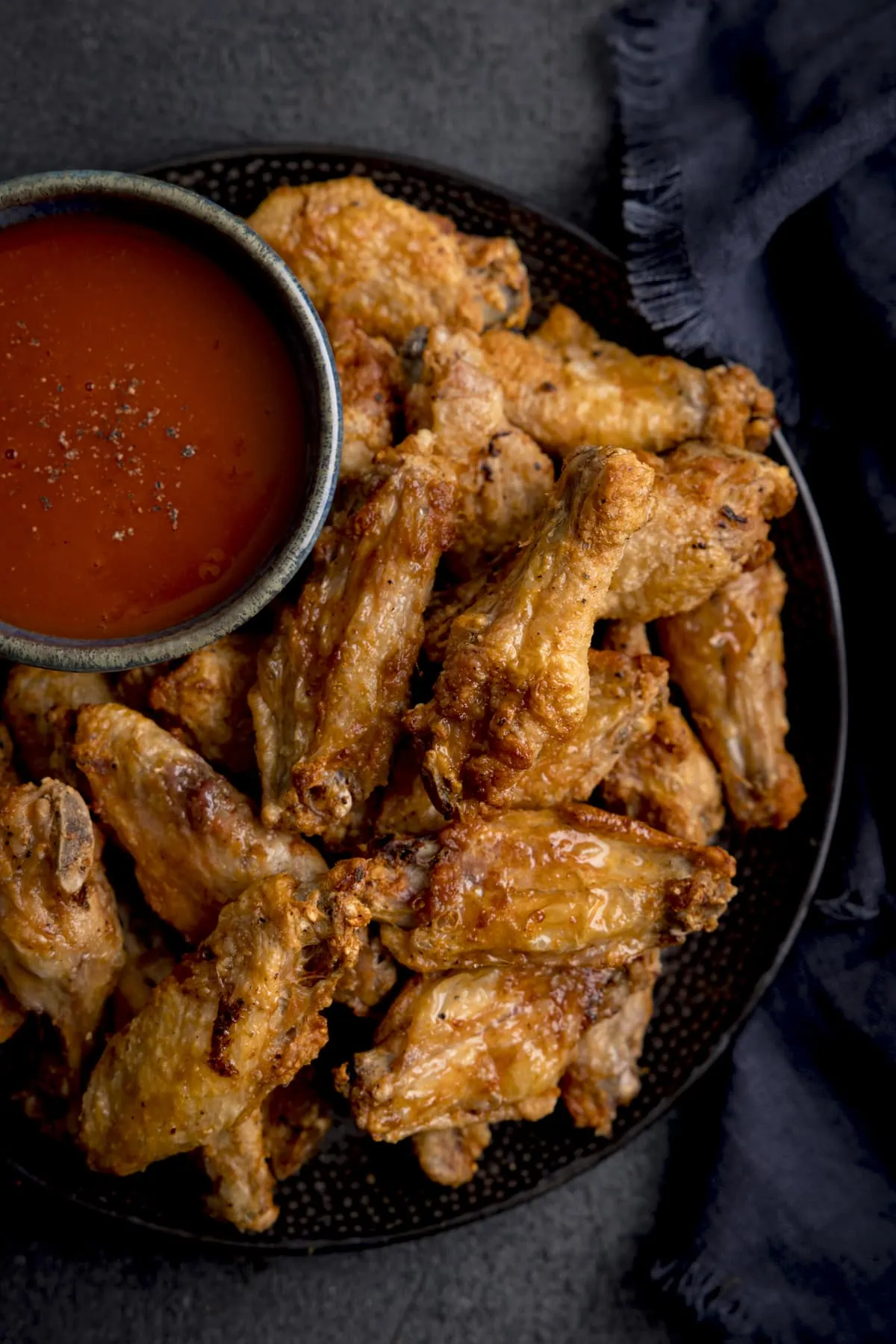 A tall shot of Air Fryer Crispy Chicken Wings piled up on a black speckled plate, thier is also a large black bowl filled with buffalo sauce, sprinkled with seasoning on the plate, the plate is on a mottled dark grey back ground and there is a large dark blue napkin to the side of the plate.