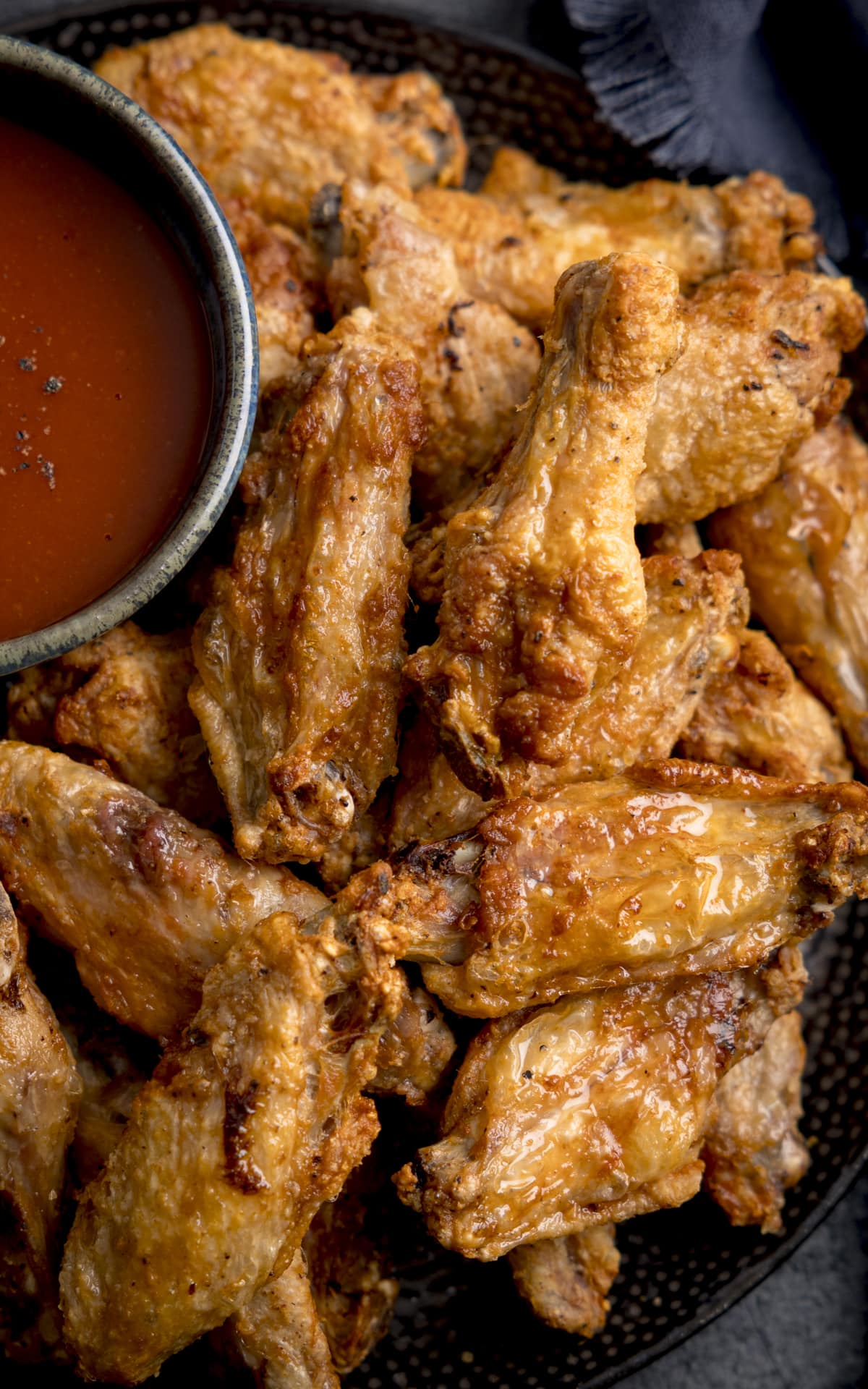 A high closeup of Air Fryer Crispy Chicken Wings stacked on a black mottled plate, there is also a large black bowl of buffalo sauce sprinkled with spices on the plate, the plate is on a mottled dark gray background and there is one large dark blue napkin on the side of the plate.