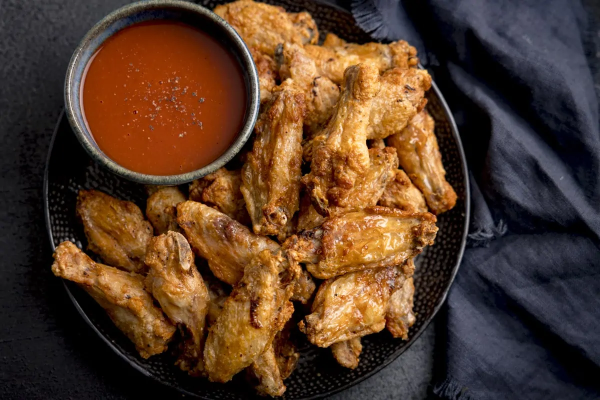 A wide overhead shot of Air Fryer Crispy Chicken Wings piled up on a black speckled plate, thier is also a large black bowl filled with buffalo sauce, sprinkled with seasoning on the plate, the plate is on a mottled dark grey back ground and there is some dark blue napkins to the side of the plate.