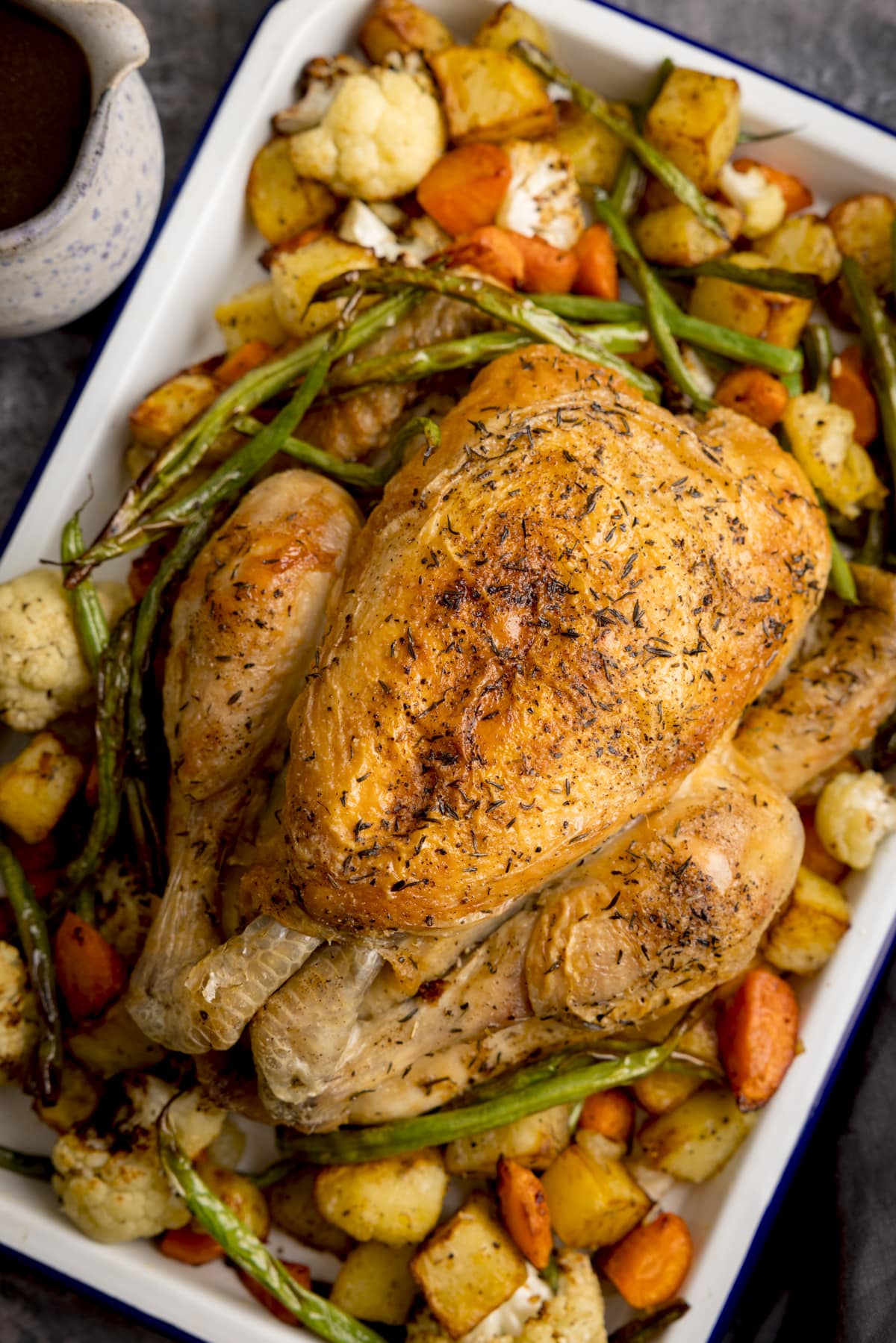 Close up overhead shot of a white roasting dish filled with Air Fryer Roast Chicken Dinner, a roast chicken in the middle with potatoes, carrots and beans surrounding it. a white jug of gravy is next to the dish and a dark blue napkin is on the side, set on a dark grey background.