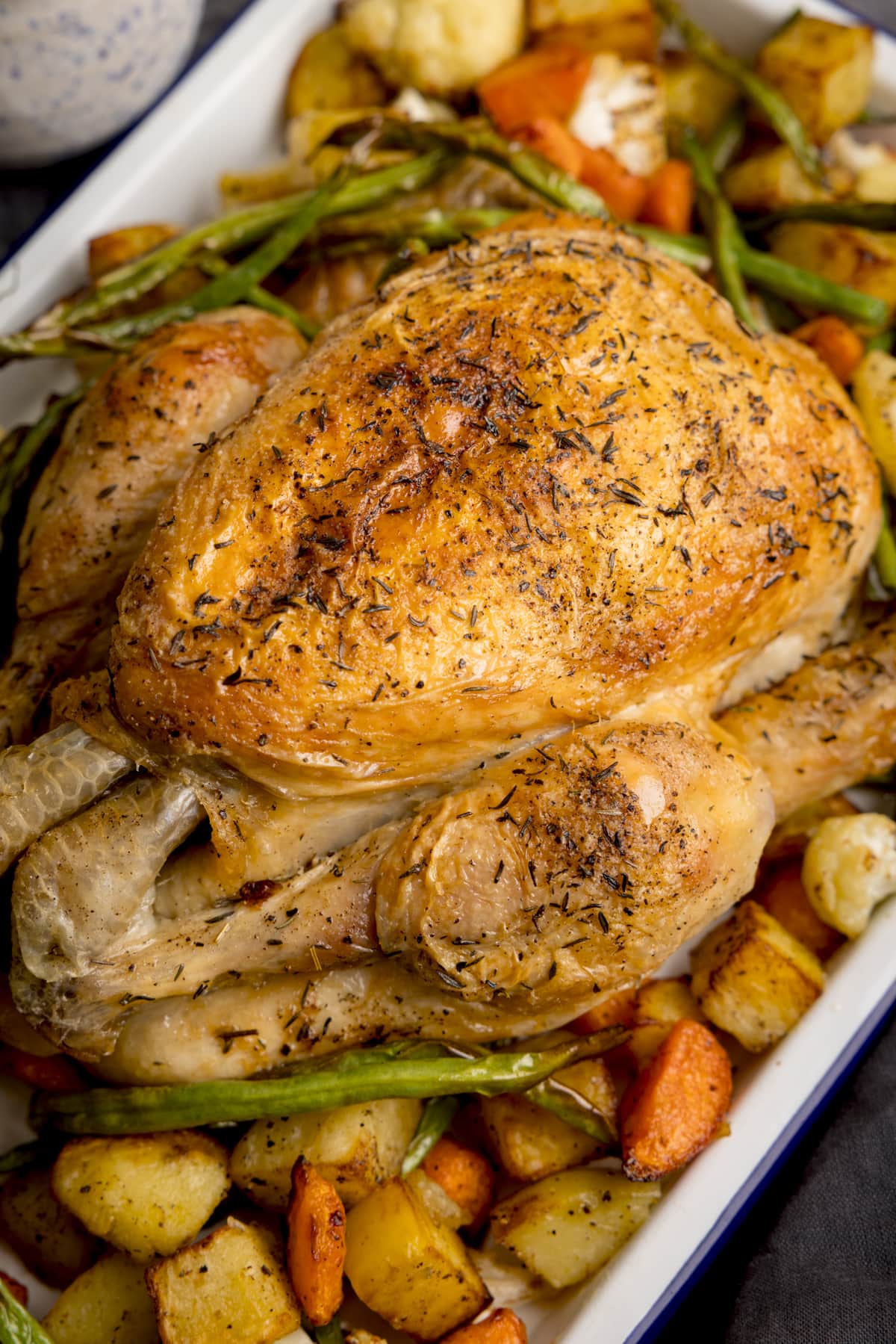 A white roasting dish filled with Air Fryer Roast Chicken Dinner, a roast chicken in the middle with potatoes, carrots and beans surrounding it. a small dark bowl filled with rock salt is next to the dish and a dark blue napkin is on the side, set on a dark grey background.