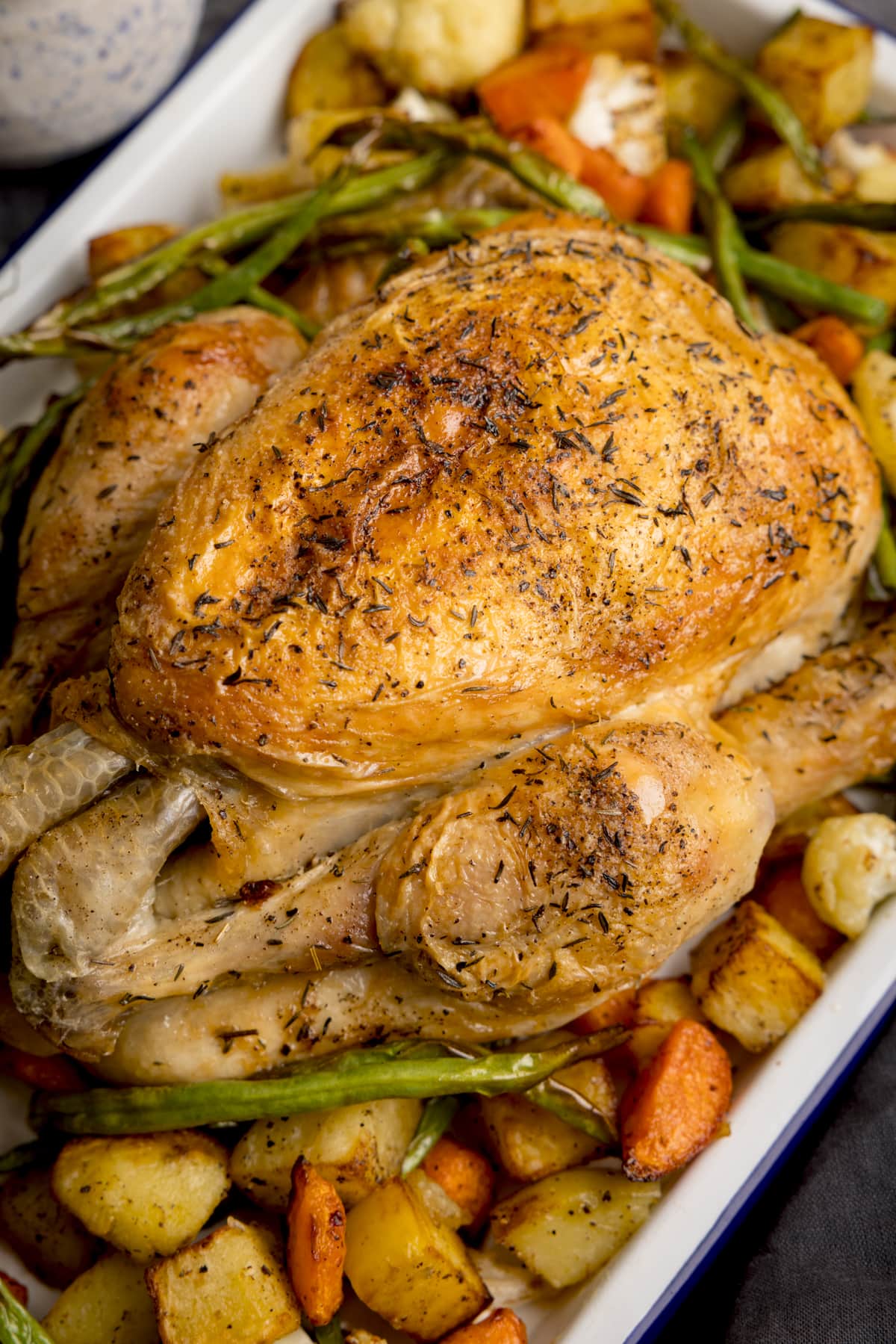 A white roaster filled with Air Fryer Roast Chicken Dinner, a roast chicken in the center surrounded by potatoes, carrots and beans. next to the plate is a small, dark bowl filled with rock salt and a dark blue serviette on a dark gray background.