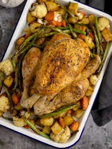 A square overhead shot of a white roasting dish filled with Air Fryer Roast Chicken Dinner, a roast chicken in the middle with potatoes, carrots and beans surrounding it. a small dark bowl filled with rock salt and a mottled blue jug filled with gravy is also next to the dish, a dark blue napkin is on the other side, set on a dark grey background.