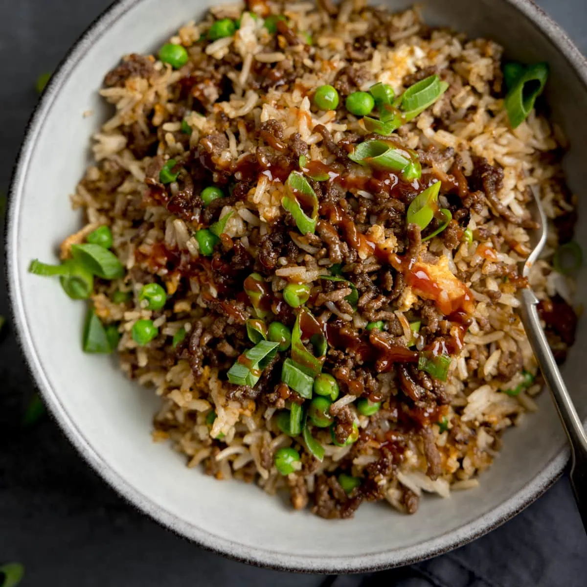 Square image of a bowl of minced beef fried rice, topped with sliced spring onions. There is a fork sticking out of the bowl.