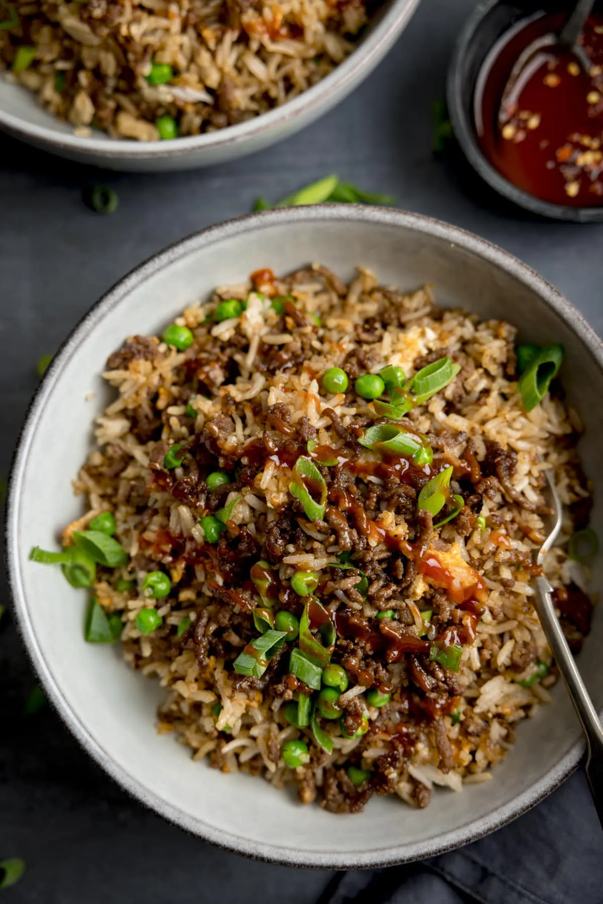 Tall image of a bowl of minced beef fried rice, topped with sliced spring onions on a dark background. There is a fork sticking out of the bowl. A second bowl of rice and a bowl of sweet chilli sauce is just in shot at the top of the frame.