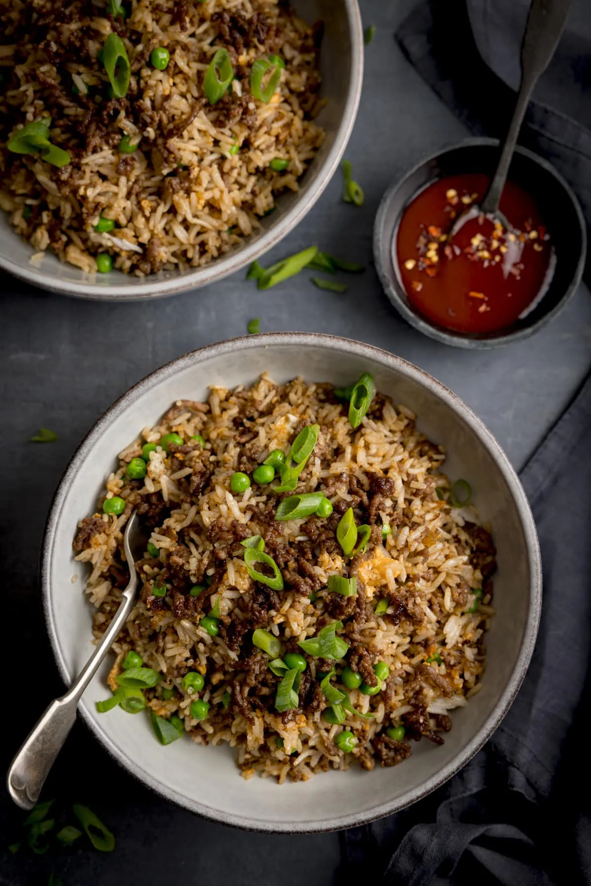 Overheat image of a bowl of minced beef fried rice, with a fork sticking out, on a dark background. A further bowl of rice and a small bowl of chilli sauce are also in shot in the top half of the frame.