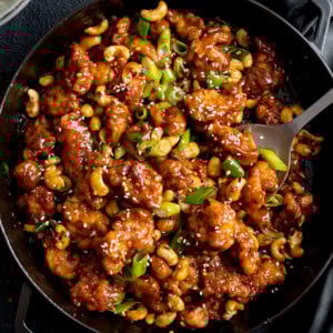A pan filled with Cashew Chicken in a glossy brown sauce, garnished with sliced spring onions and sesame seeds, with a serving spoon on the side.