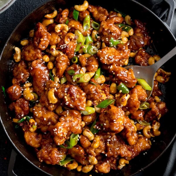 A pan filled with Cashew Chicken in a glossy brown sauce, garnished with sliced spring onions and sesame seeds, with a serving spoon on the side.
