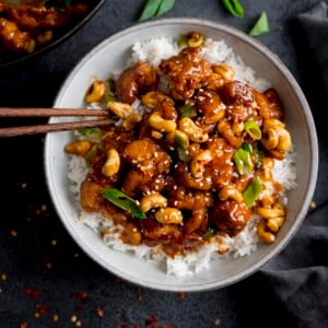 Square overhead image of sticky cashew chicken with rice in a white bowl on a dark background. There are chopsticks sticking out of the bowl.