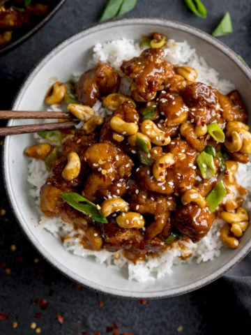 Square overhead image of sticky cashew chicken with rice in a white bowl on a dark background. There are chopsticks sticking out of the bowl.