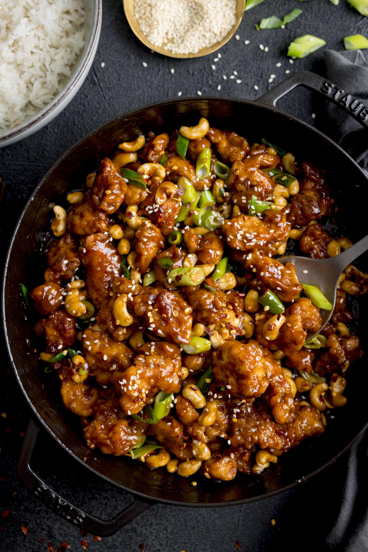 Overhead of crispy cashew chicken with spring onions in a black pan. There is a bowl of rice and small bowl of sesame seeds in shot.